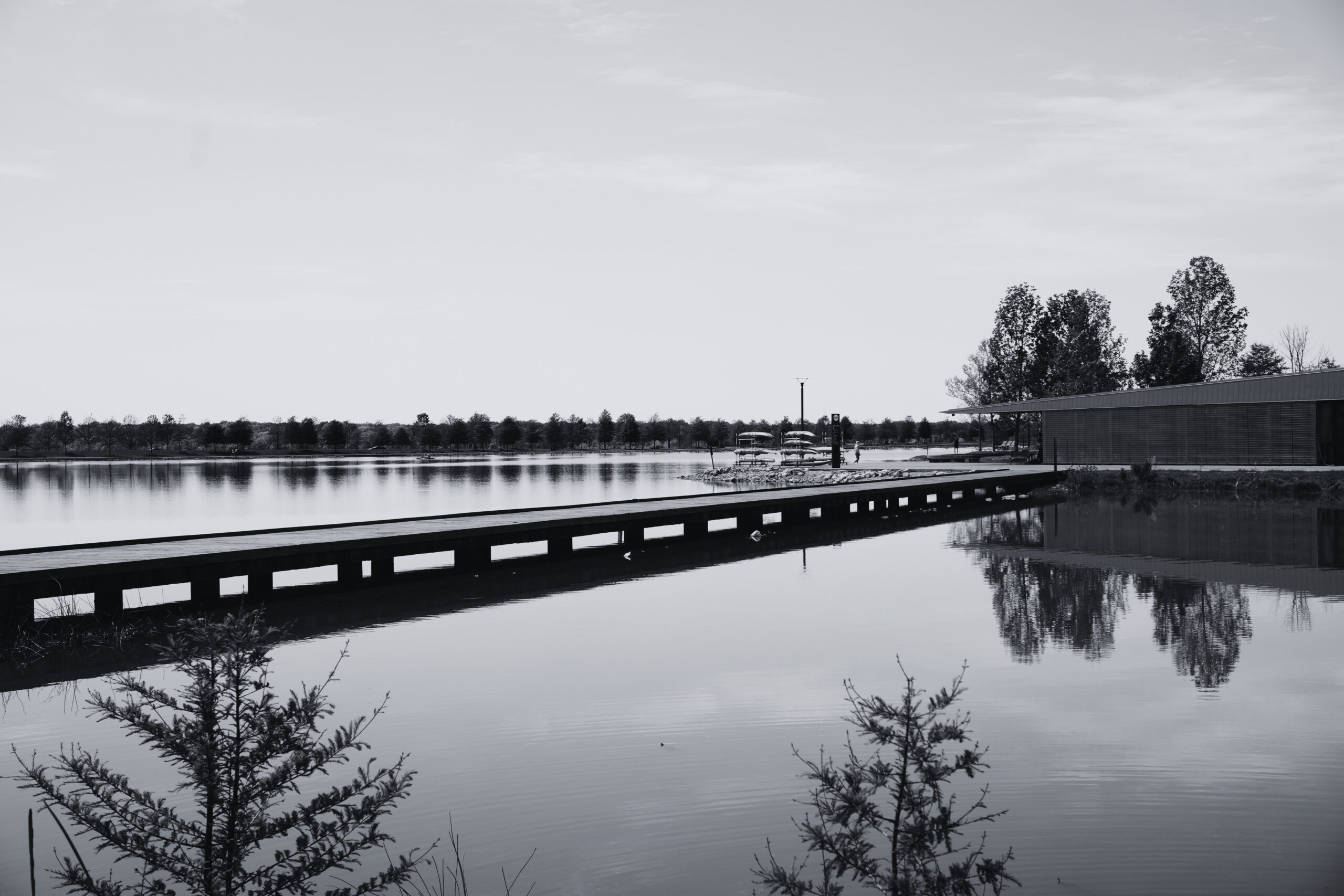 Grayscale landscape: a calm lake with a wooden boardwalk across, a low rectangular building on the right, and trees along the horizon behind.