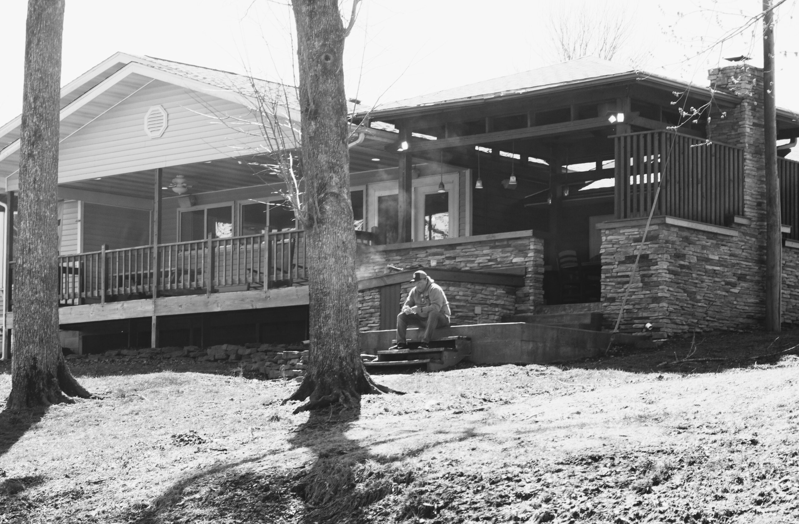 Man seated on concrete steps in front of a two-level house with a wraparound porch, stone veneer, and surrounding trees (black-and-white photo).